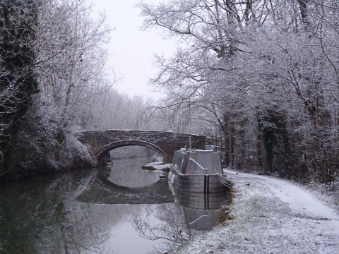 A silver narrowboat moored against a snowy canal bank with an arched bridge behind it.