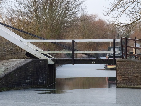 Thrupp, 11 Feb A lift bridge over a canal with ice on each side and a strip of clear water beneath it. Behind the bridge there are trees on the left bank and the stern of a moored boat on the right.