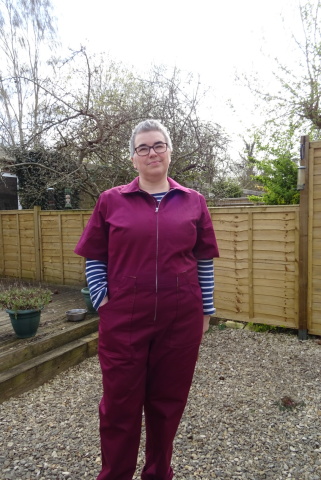 a white woman with short hair and glasses stands in a garden, wearing a dark red boiler suit over a blue and white striped t-shirt.
