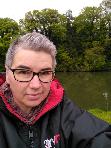 A selfie of a white person with short grey-brown hair and glasses, wearing a black top with a hood lined in red fleece, with a river and trees behind her.