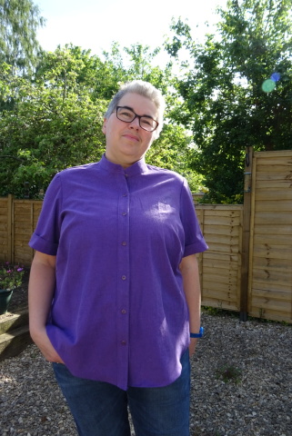 Purple shirt A white person with short grey hair and glasses stands in a garden wearing a purple collarless shirt with short sleeves.