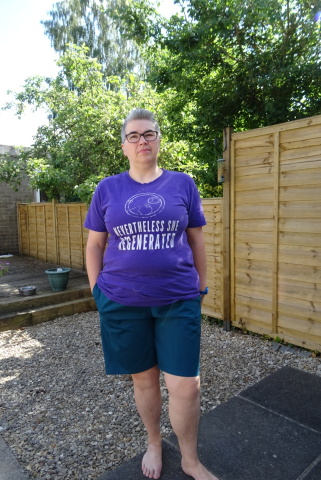 A white person with short grey hair and glasses stands in a garden wearing a purple t-shirt with white printing and a pair of dark teal knee-length shorts.