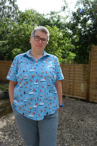 A white person with short grey hair and glasses stands in a garden wearing a pale blue short-sleeved collared shirt with a print of people swimming.