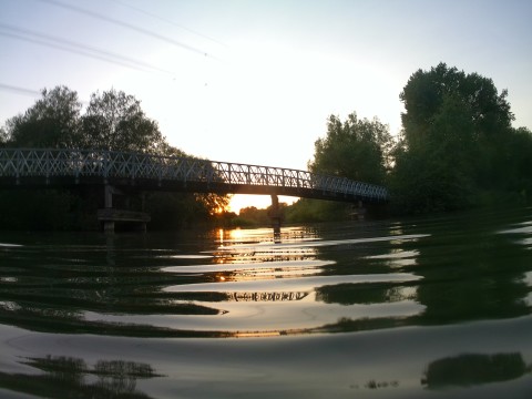 The setting sun shining under a footbridge between tree-lined river banks.