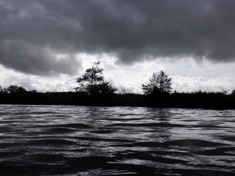 Dark clouds and grey water with the riverbank and two trees silhouetted against a line of brightness in between.