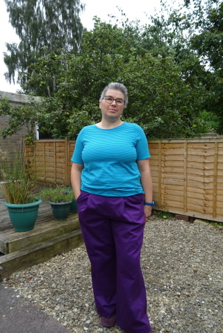 Harper Pants A white person with short grey hair and glasses stands in a garden wearing a teal and turquoise striped top and wide-legged violet trousers.