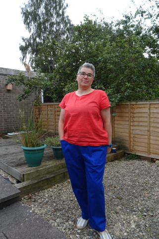 Pietra Pants A white person with short grey hair and glasses stands in a garden wearing a red top and bright blue trousers.