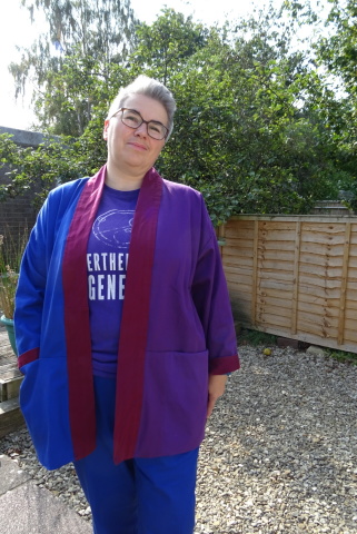 Jacket! A white person with short grey hair and glasses stands in a garden wearing an oversized jacket which is bright purple on one side and cobalt blue on the other with a dark red collar over a purple t-shirt with white writing and cobalt blue trousers.