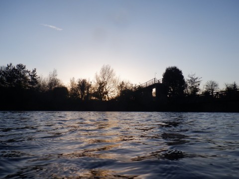 A lake reflecting deep blue sky and orange sunset light, with trees and a metal footbridge silhouetted against the sky behind the water.