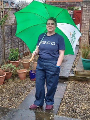 A white person with short grey hair and glasses stands in a rainy garden holding a large green and white umbrella. They're wearing a navy t-shirt with DISCO printed on it in white and dark denim trousers with large pockets and a button fly.